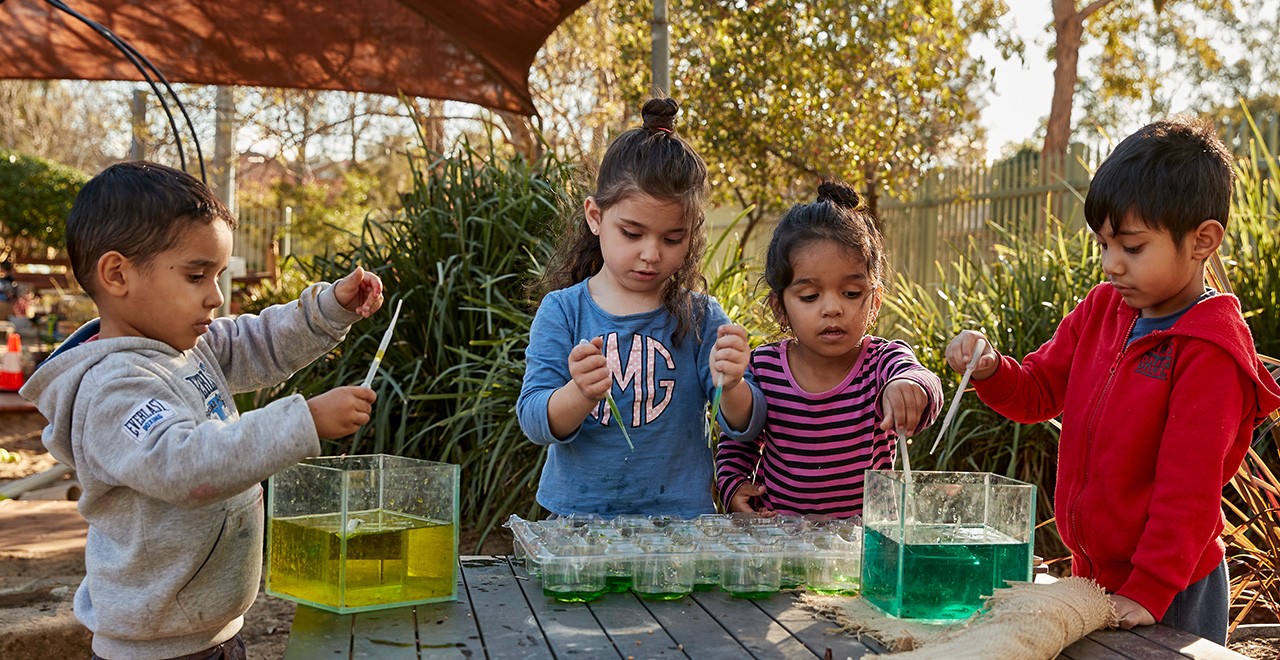 Four children playing outside at a table mixing different coloured liquids in clear plastic contains of various size