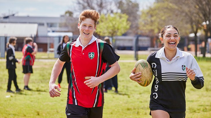A male and female high school student running together on the school oval with a rugby ball.